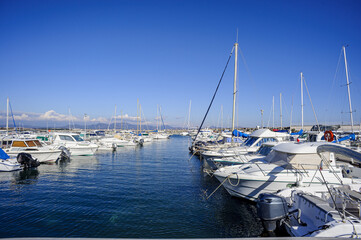Pleasure boats moored in a small port on the Mediterranean coast. The water reflects the blue of the sky. In the background, the Esterel mountains stand out on the horizon.