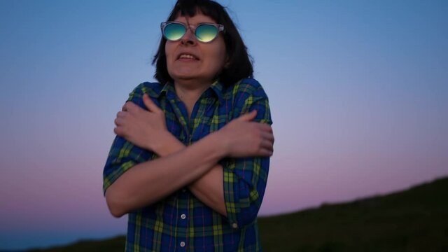 Slow motion young woman in mirrored sunglasses hugging herself crossed her hands and rubbing her shoulders because of feeling freeze on hill at dusk after sunset.Female teeth are chattering from cold