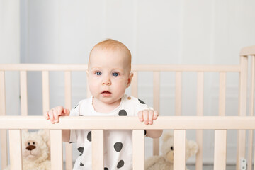 portrait of a baby 8 months old standing in a crib with toys in pajamas in a bright children's room after sleeping and looking at the camera, a place for text