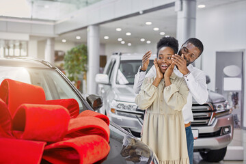 young and beautiful married couple in cars showroom, confident black man buy new car as a present for his wife - obrazy, fototapety, plakaty