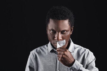 Sad African-American man removing tape from mouth on dark background. Stop racism