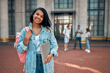 Cute African American girl student with a backpack and laptop near the campus against the background of a group of students.