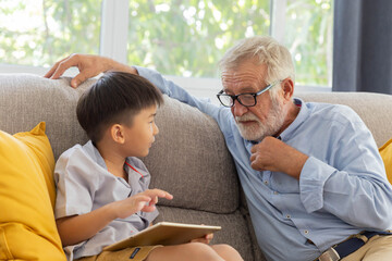 Obraz premium Grandfather and little cute grandson playing touchscreen tablet PC together on sofa in their house