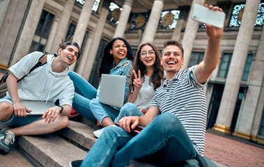 A group of students are sitting on the steps near the campus with laptops, relaxing, chatting and...
