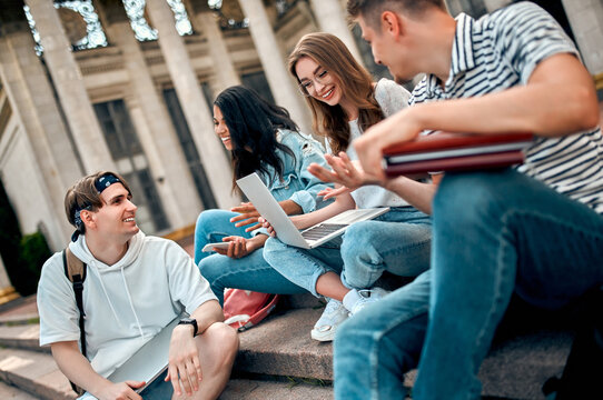 A group of students with laptops sit on the steps near the campus and communicate.