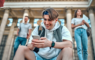A student guy with a backpack sits on the steps near the campus and uses his smartphone in the background of students passing by.