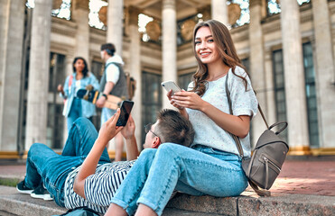 A couple of students with backpacks and a laptop sit on the steps outside the campus and use their smartphones.