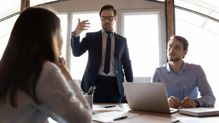 Motivated caucasian male team leader stand talking brainstorming at office meeting with colleagues, confident businessman boss lead briefing with diverse employees, cooperation, leadership concept