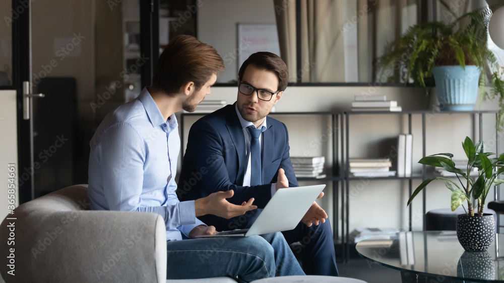 Poster concentrated caucasian male employees sit on couch in office brainstorm work together on laptop, foc