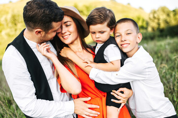 Midshot of a happy family outside on nature. Shot of a loving family. Young girl hugs and touches her husband while holding a little boy, the older son is hugging mom.