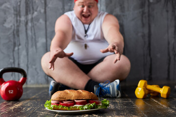 obese young guy going to eat big sandwich despite recent sport training, se sits on the floor with...