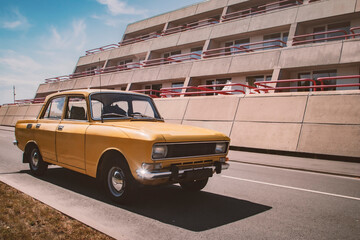 A nostagic vintage photo of a yellow retro classic soviet russian AZLK Moskvich 2140 car wearing an old black blank licence plate parked in front of a 1980s concrete building on a sunny summer day - obrazy, fototapety, plakaty