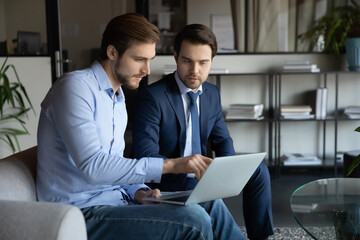 Serious Caucasian businessmen sit on couch in office look at laptop screen discussing business project together, male partners colleagues brainstorm work together on computer, cooperation concept