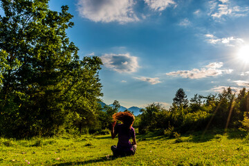Fototapeta na wymiar A full body shot of an unrecognizable young Caucasian redhead woman sitting on the grass in a meadow in a forest in the French Alps during sunset