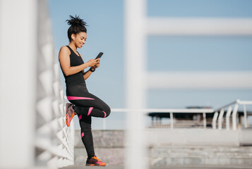 Obraz premium African american girl in sport uniform leans on wall with wireless headphones and looks at phone