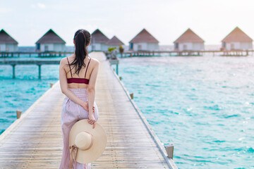 Beautiful young girl walking smiling resting relaxing on the tropical beach. Young girl standing on wooden jetty wearing swimwear in sunrise. Woman with hat by beautiful blue ocean, turquoise water.