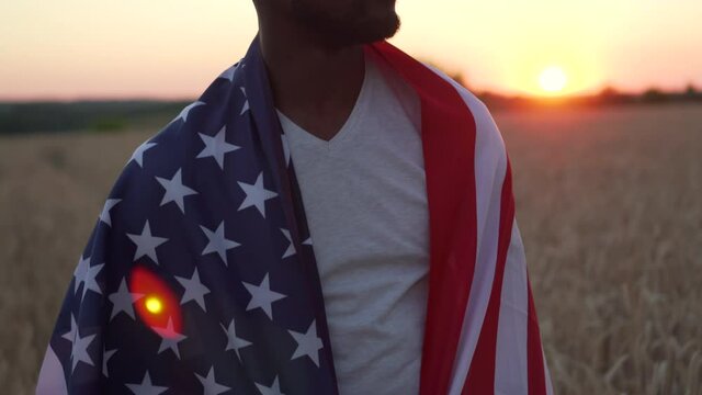 Portrait of an afro american man with the national flag of usa on his shoulders in a field at sunset. Flare effect.