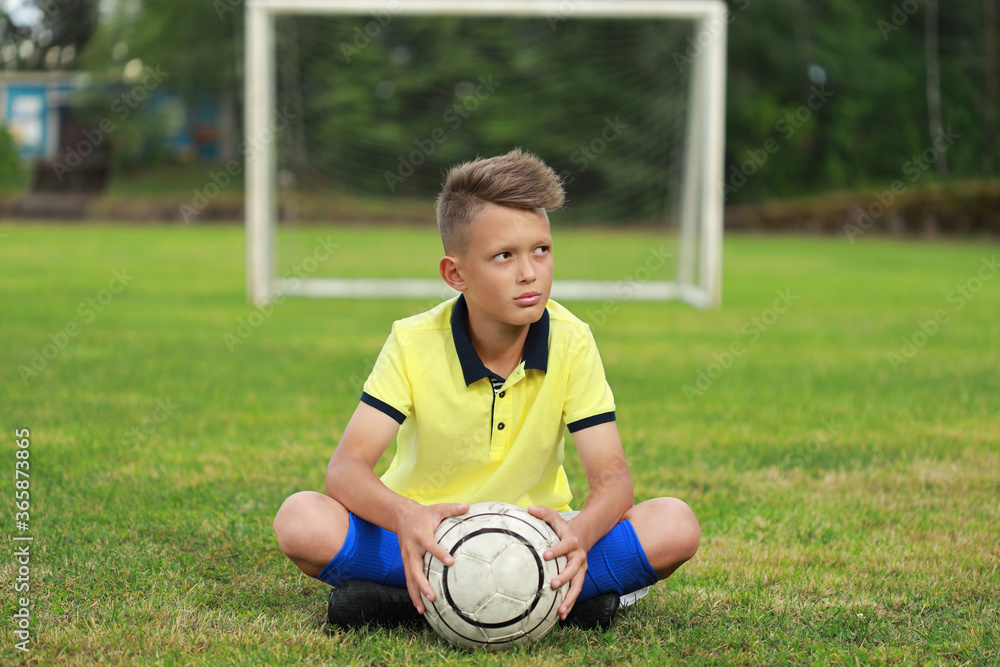 Poster handsome boy soccer player sits on the soccer field against the backdrop of the goal with the ball