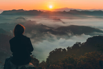 Woman tourist is sitting on a rocky mountain, looking at the beautiful of the sunrise and the fog covered the mountains in the morning, to travel concept.