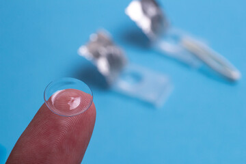 tweezers hold a one-day contact lens against a blurry background of glasses and lens cases