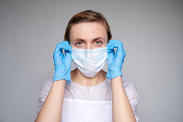 Close up of female doctor or scientist with a medical mask and hands in latex gloves over grey background. She is adjusting a mask with her hands. - Powered by Adobe