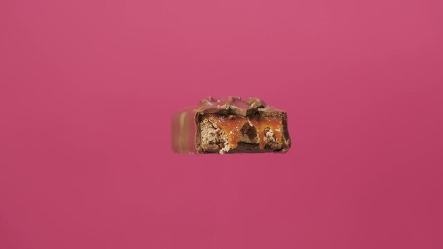 A delicious and unhealthy chocolate bar rotates on a pink background. Bitten sweets isolated. The unpacked bar is spinning. Nougat and caramel inside chocolate