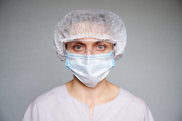 Close up of female doctor or scientist with a medical mask and surgical cap and glasses over grey background.