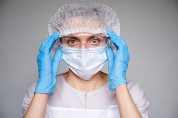 Close up of female doctor or scientist with a medical mask, surgical cap, glasses and hands in gloves over grey background.