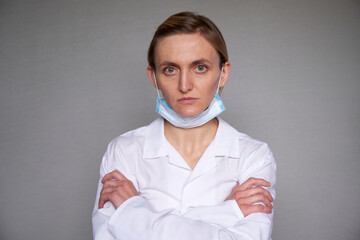 Close up of female doctor or scientist in protective facial mask over grey background. Medical mask is lowered to the chin. Arms in crossed over chest