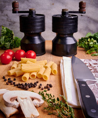 Pasta ingredients on chopping board with salt and pepper grinders and kitchen knife.