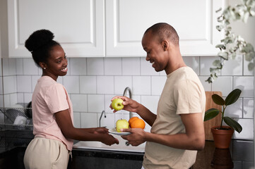 kind careful man with wife in the kitchen, beautiful young lady is washing fruits while man stands with plate, they have conversation and smile