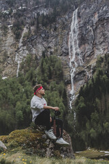 Obraz premium Obersee. Schonau am Konigssee. Bavaria. Germany. Young hipster photographer with camera sits on the stone & looks at Rothbach waterfall on background of pine forest & rock mountain cliff
