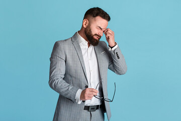 Stressed busnessman crying over mistake or suffering from headache on blue background