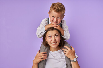 portrait of cute mother and kid boy isolated over purple background, awesome child sit on mom's neck, woman and chil in love, happy together