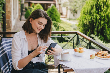 Young woman using her smart phone during breakfast on cozy terrace of a country house.