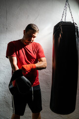 Boxeador joven principiante con camiseta roja poniendose cinta en las manos junto a un saco negro...