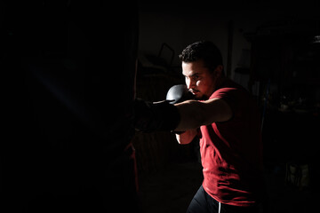 Fototapeta na wymiar Boxeador joven principiante con camiseta roja golpeando un saco negro en su garaje con guantes de boxeo negros, iluminado con luz intensa de flash y fondo negro