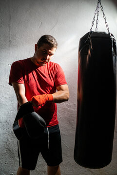 Boxeador joven principiante con camiseta roja poniendose cinta en las manos junto a un saco negro en su garaje con guantes de boxeo negros, iluminado con luz intensa de flash y fondo negro