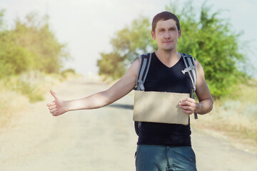 Hitch-hiking traveler with blank cardboard and raised thumb up on the road while hitchhiking. Budget travel. Copyspace