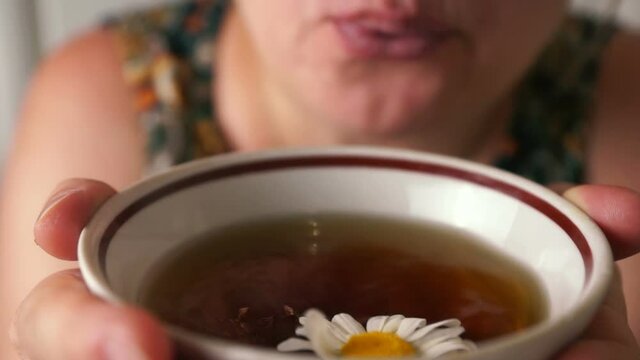 Close-up of lips of a Caucasian woman blowing out fresh chamomile tea in a cup she holds in her hands. A decoction of chamomile for colds