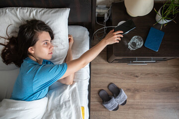 awake woman taking phone from wireless charger from bedside table - obrazy, fototapety, plakaty