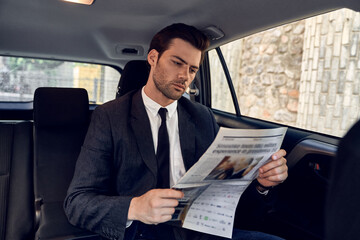 Reading latest news. Handsome young man in full suit reading a newspaper while sitting in the car
