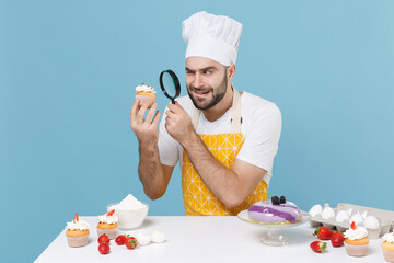 Pensive bearded male chef or cook baker man in apron white t-shirt toque chefs hat cooking at table isolated on blue background. Cooking food concept. Looking on dessert cupcake with magnifying glass.
