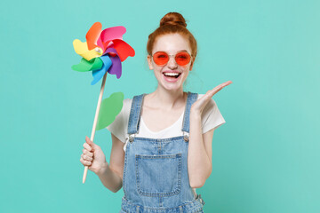 Funny young readhead girl in casual denim clothes eyeglasses posing isolated on blue turquoise wall background studio. People lifestyle concept. Mock up copy space. Hold toy windmill, spreading hands.