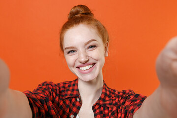 Close up of smiling young readhead girl in casual red checkered shirt posing isolated on orange wall background studio. People lifestyle concept. Mock up copy space. Doing selfie shot on mobile phone.