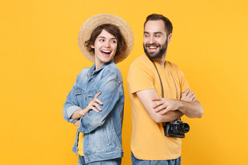 Obraz premium Cheerful tourists couple friends guy girl in summer clothes hat with photo camera isolated on yellow background. Passenger traveling abroad on weekends. Air flight journey concept. Stand back to back.