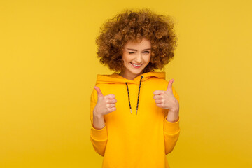 Good job! Portrait of optimistic curly-haired young woman in urban style hoodie winking and showing...