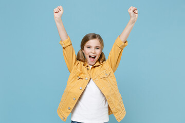 Excited little blonde kid girl 12-13 years old in yellow jacket posing isolated on blue wall background children studio portrait. Childhood lifestyle concept. Mock up copy space. Doing winner gesture.
