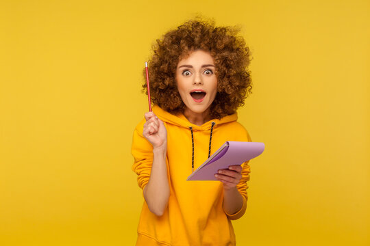 Portrait Of Inspired Joyful Curly-haired Woman In Urban Style Hoodie Looking Amazed With Sudden Genius Idea And Holding Pencil To Write In Notebook. Indoor Studio Shot Isolated On Yellow Background