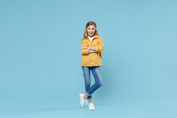 Full length portrait of pretty little blonde kid girl 12-13 years old in yellow jacket isolated on blue wall background studio. Childhood lifestyle concept. Mock up copy space. Holding hands crossed.